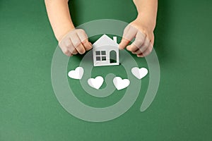 Family home and real estate concept. Close up of child hands holding white paper house and heart on green background.