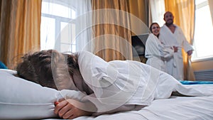 A family in the hotel room - a little girl sleeping in bed - her mom and dad looking at her and opening the curtains