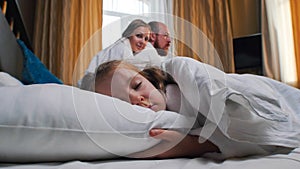 A family in the hotel room - a little girl sleeping in bed - smiling mom and dad looking at her
