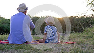 Family idyll, daddy with son in straw hats rest on blanket on background of sunset at rural in open air