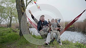 Family, joyful little grandson is sitting on hands of caring and joyful grandad loving grandmother in hammock and