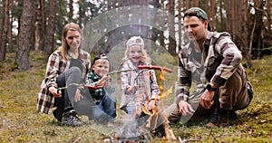 family with kids cook sausages on wooden skewers over a campfire in the forest. nature picnic, outdoor activities