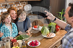 Family In Kitchen Cooking Food Father Taking Photo On Cell Smart Phone Of Mother Son And Daughter Sitting At Table