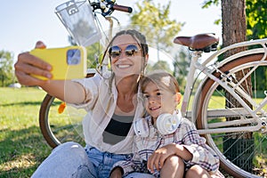 family, leisure and technology concept - happy mother and little daughter with smartphone and bicycles in summer park