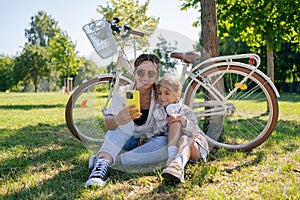 family, leisure and technology concept - happy mother and little daughter with smartphone and bicycles in summer park