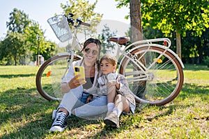 family, leisure and technology concept - happy mother and little daughter with smartphone and bicycles in summer park