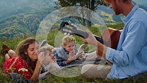 Family listening guitar music on green hill. Father playing for woman with kids.