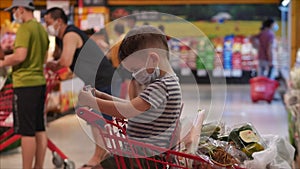 Family makes purchases in hypermarket, child in a protective mask from viruses sits in a grocery cart watches videos on