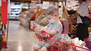 Family makes purchases in hypermarket, child in a protective mask from viruses sits in a grocery cart watches videos on