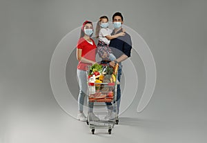 Family in medical masks with shopping cart full of groceries on light grey background