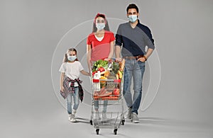 Family in medical masks with shopping cart full of groceries on light grey background