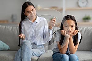 Family misunderstanding concept. Offended asian daughter sitting on couch, turning back to mother after quarrel