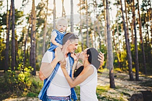 Family mom, dad and daughter sits at the daddy on the shoulders, and parents kiss on the nature in the forest in the summer at sun