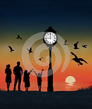 A family, mom, dad and kids are seen with a flock of seagulls on the beach where a lighted clock is seen at sunSet