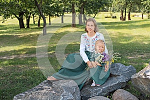 Family mom with daughter in vintage retro linen dresses sitting on a stone rock in the park forest with bouquet of flowers