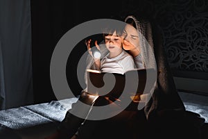 Family Mom and little son reading a book with a flashlight under the blanket