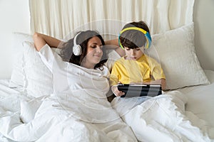 Family morning in bedroom: mom in headphones look at little kid son play games on tablet computer