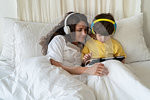 Family morning in bedroom: mom in headphones look at little kid son play games on tablet computer