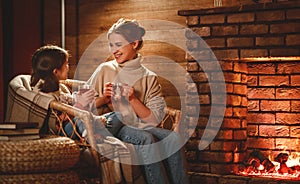 Family mother and child reading book and drink tea on winter evening by fireplace