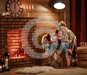 Family mother and child reading book and drink tea on winter evening by fireplace