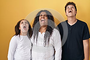 Family of mother, daughter and son standing over yellow background angry and mad screaming frustrated and furious, shouting with