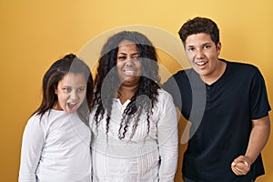 Family of mother, daughter and son standing over yellow background angry and mad screaming frustrated and furious, shouting with