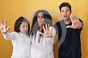 Family of mother, daughter and son standing over yellow background doing stop gesture with hands palms, angry and frustration