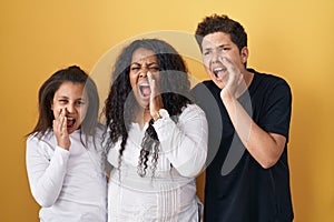 Family of mother, daughter and son standing over yellow background shouting and screaming loud to side with hand on mouth