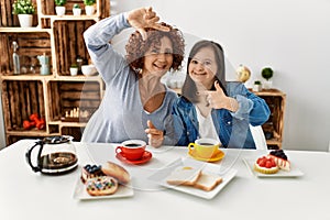Family of mother and down syndrome daughter sitting at home eating breakfast smiling making frame with hands and fingers with
