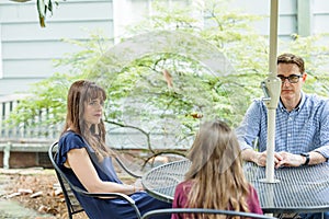 A family of a mother and father and two daughters sitting outside at a patio table with an umbrella
