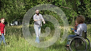 Family with a mother in wheelchair playing with children outdoors throwing ball and frisbee. Mother and father with