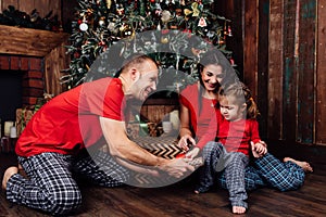 Family in pajamas plays next to a Christmas tree and a fireplace. A little daughter takes a gift from her father.