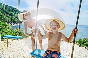 Family photo of smiling mom and happy son having fun together on the swing, enjoying sunny day on the tropical beach. Thailand.