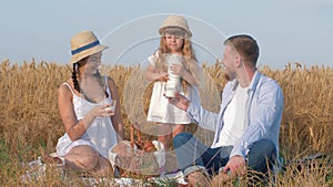 Family picnic in grain field, little kid girl pours milk into the glasses of her young cheerful parents during having