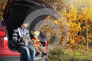 Family picnic outdoor, road trip in autumn season. Father and his little child sitting inside car trunk near rural road in autumn