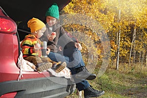 Family picnic outdoor, road trip in autumn season. Mother and her little child sitting inside car trunk near rural road in autumn