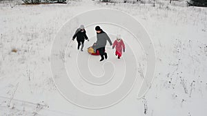 Family playing on the winter field - father rides his son on the inflatable sled and his wife and daughter running near