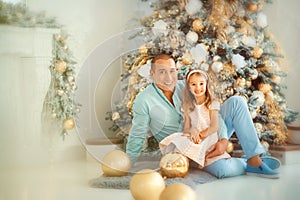 Family portrait of happy father in blue clothes and his 4 years old daughter sitting on his lap near christmas tree. New year and