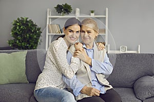 Family portrait of mature mother and her adult daughter hugging while sitting on sofa at home.