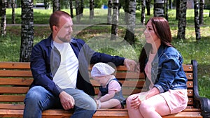 Family portrait mom, dad sitting talking on bench in city park with baby girl.