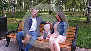 Family portrait mom, dad sitting talking on bench in city park with baby girl.