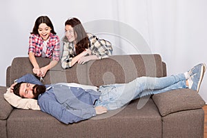 Family prank. Mom and daughter going to cut beard sleeping dad. Lets have fun. Cheerful girls with scissors cut long