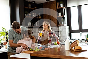 Family prepares a meal together, with dad helping daughter cut vegetables and mom enjoying a juice