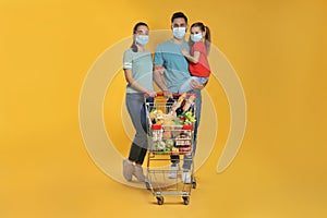 Family with protective masks and shopping cart full of groceries on yellow background