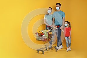 Family with protective masks and shopping cart full of groceries on yellow background. Space for text