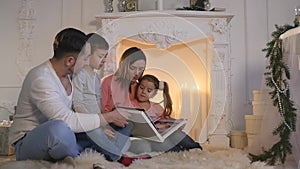 Family read book sitting on sofa in front of fireplace in Christmas decorated house interior