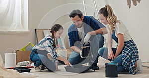 Family is renovating new house. Dad in shirt pours brown paint from bucket in cuvette. Daughter with pigtails and mother