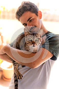 Vertical photo. Young man hugs a green-eyed tabby cat. The cat looks at the camera.