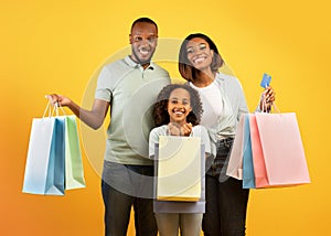 Family shopping. Excited african american parents and their cute daughter holding shopper bags over yellow background
