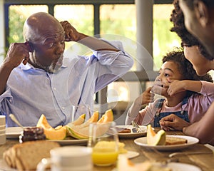 Family Shot With Grandparents Parents And Granddaughter Pulling Faces At Breakfast Around Table At Home